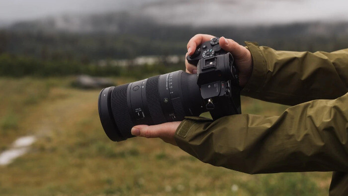 Фотограф у куртці тримає камеру Sony з об'єктивом Sigma 28-105mm f/2.8 DG DN Art двома руками на фоні туманного гірського пейзажу
